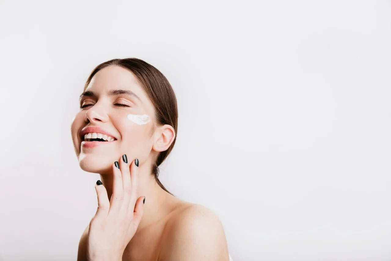 Mulher sorrindo com creme facial aplicado na bochecha, sobre fundo branco